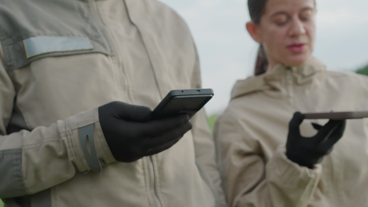 close up adult wearing protective gloves holding smartphone while companion gestures to sky in lush green field, subtle interaction and focus on device controls with soft background blur