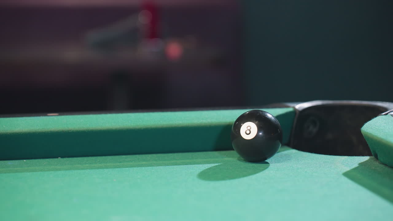 Close-up of black billiard ball rolling toward pool pocket after being struck by white cue ball on green felt table. Focus on precision and intensity of gameplay with dramatic lighting and shadows