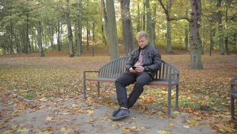A-young-attractive-male-sitting-on-a-park-bench-using-his-smart-phone-on-an-autumn-day