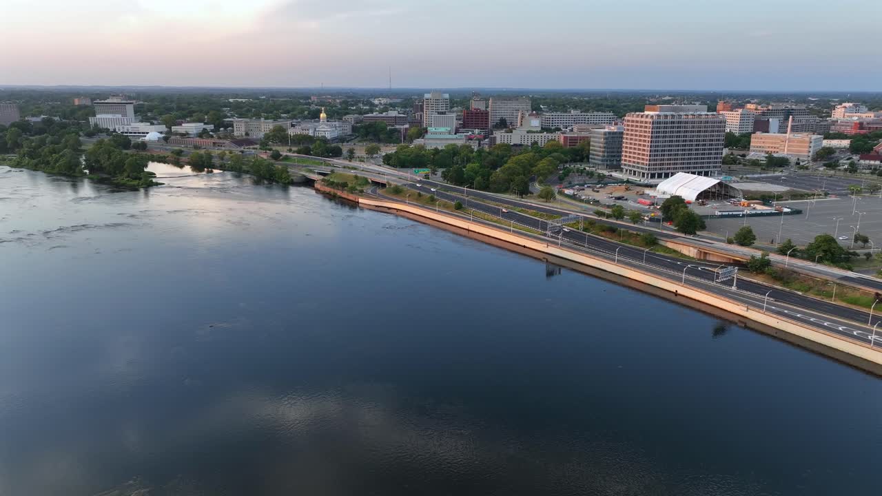 Trenton cityscape near River during golden sunrise. Aerial wide shot. Traffic on shoreline road. Apartment blocks and offices in American city downtown