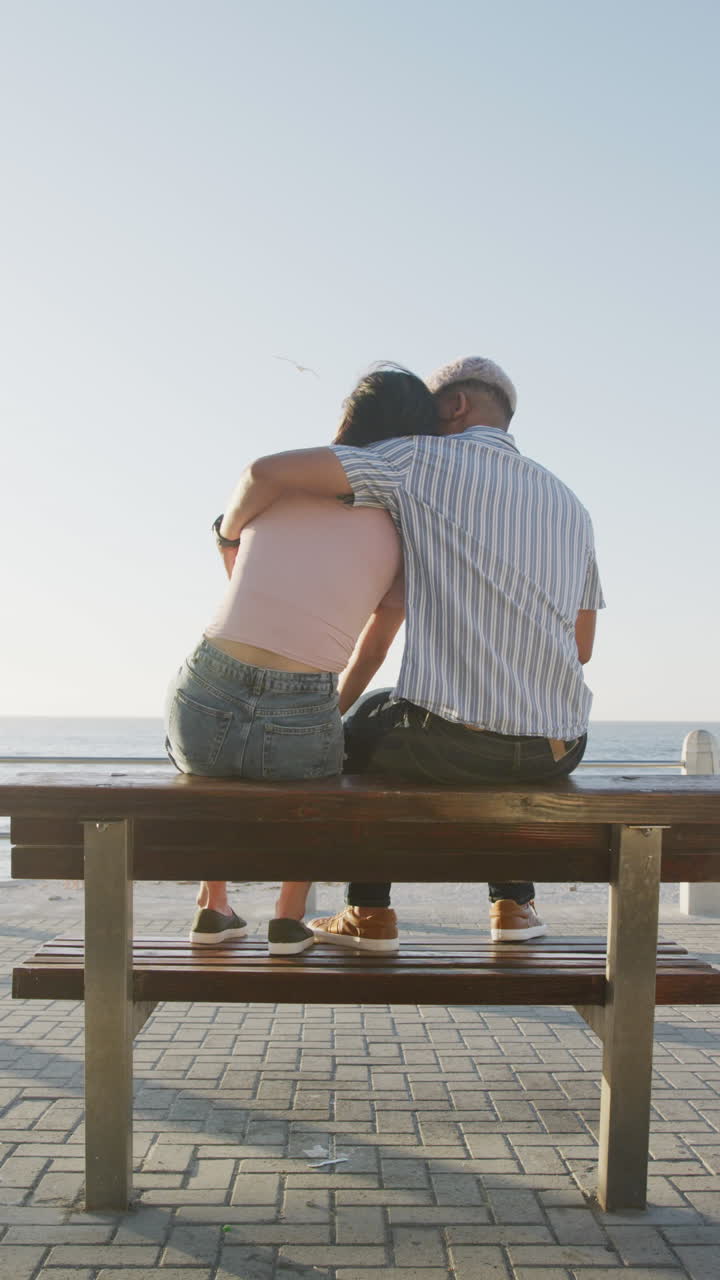 Vertical video of happy biracial couple sitting on bench and embracing on promenade, in slow motion