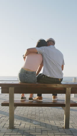 vertical video of happy biracial couple sitting on bench and embracing on promenade, in slow motion