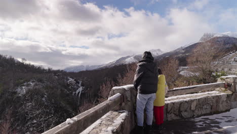 mother and child admire the vuliñanco waterfall from a snowy stone lookout on route 40, surrounded by patagonian mountains and wintry forest landscape, static camera, real time