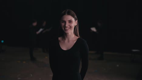 retrato de una joven actriz confiada en un traje negro entre los actores que se están preparando para una actuación y ensayando en el escenario en un teatro con cortinas negras