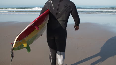 vista trasera de un surfista masculino con una pierna biónica yendo al océano con una tabla de surf bajo el brazo