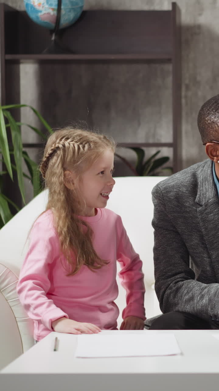 Happy girl student raises hand to answer while confused boy thinks at English lesson with positive black man tutor at private lesson at home slow motion