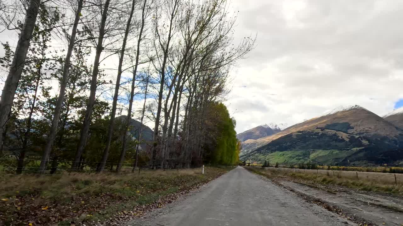 Vehicle travels rural gravel road lined with trees, mountains visible, overcast daylight, steady forward motion