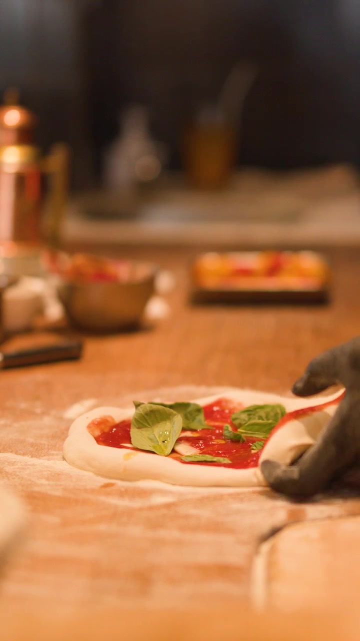 Primo piano di mani guantate che preparano una pizza con salsa di pomodoro e basilico su un bancone di legno