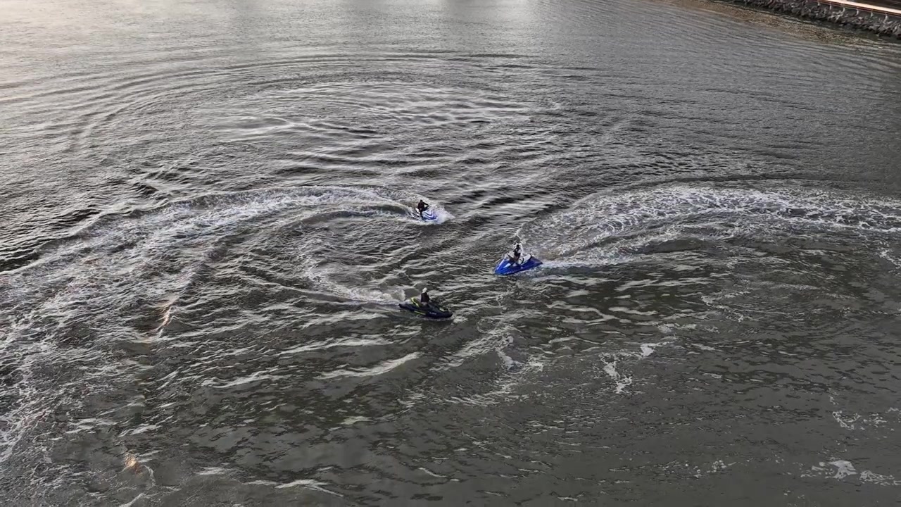 An aerial view over the Spuyten Duyvil Creek on a cloudy day. There are three people on jet skis floating in the creek. The camera is tilt down over the trio and is stationary, filming in slow motion.