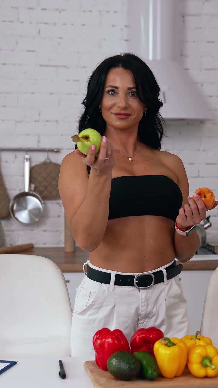 Beautiful woman in her kitchen holding fruit in both hands. Lady wearing black top and white jeans looks at fruit in her hands and then offers a peach and an apple. Vertical video