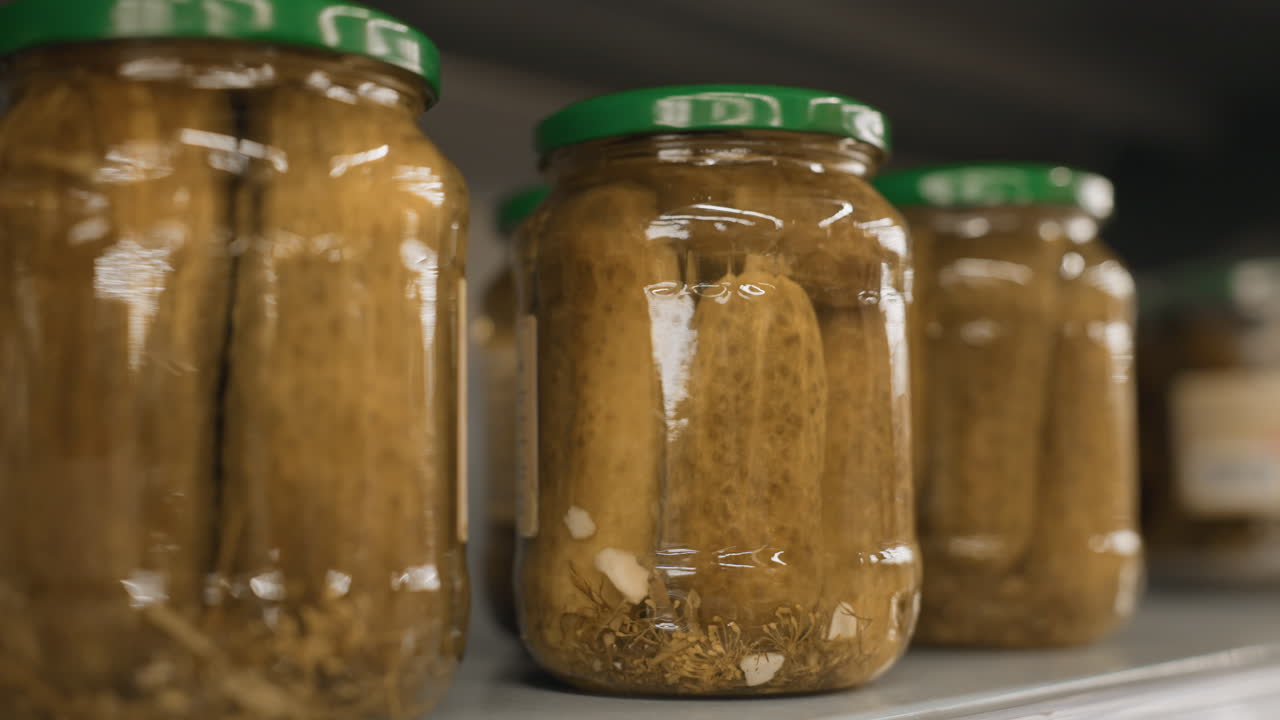 Close up of lone shopper hands tapping transparent glass jar of pickles on supermarket shelf grid highlighting polished nails and metal rails in blurred grocery aisle background retail scene