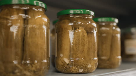 close up of lone shopper hands tapping transparent glass jar of pickles on supermarket shelf grid highlighting polished nails and metal rails in blurred grocery aisle background retail scene