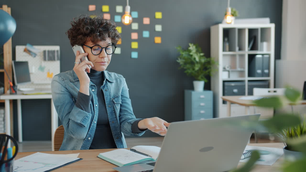 Kvinde på kontor arbejder på bærbar computer og taler i telefon