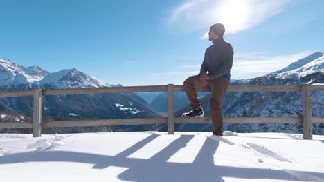 Young-caucasian-man-with-a-beanie-and-sunglasses-is-sitting-down-on-a-wooden-railing-and-enjoying-the-view-on-a-sunny-day-in-the-mountains