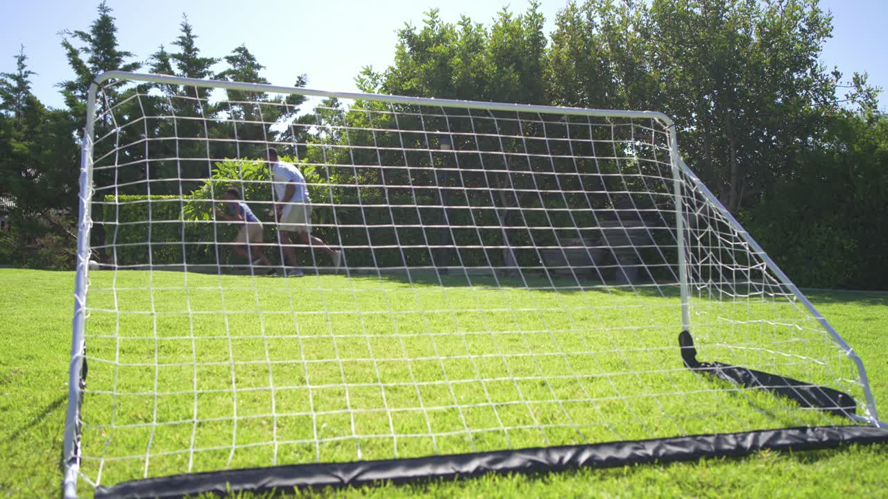 African American father guarding portable goal in backyard while teenage son dribbling and scoring