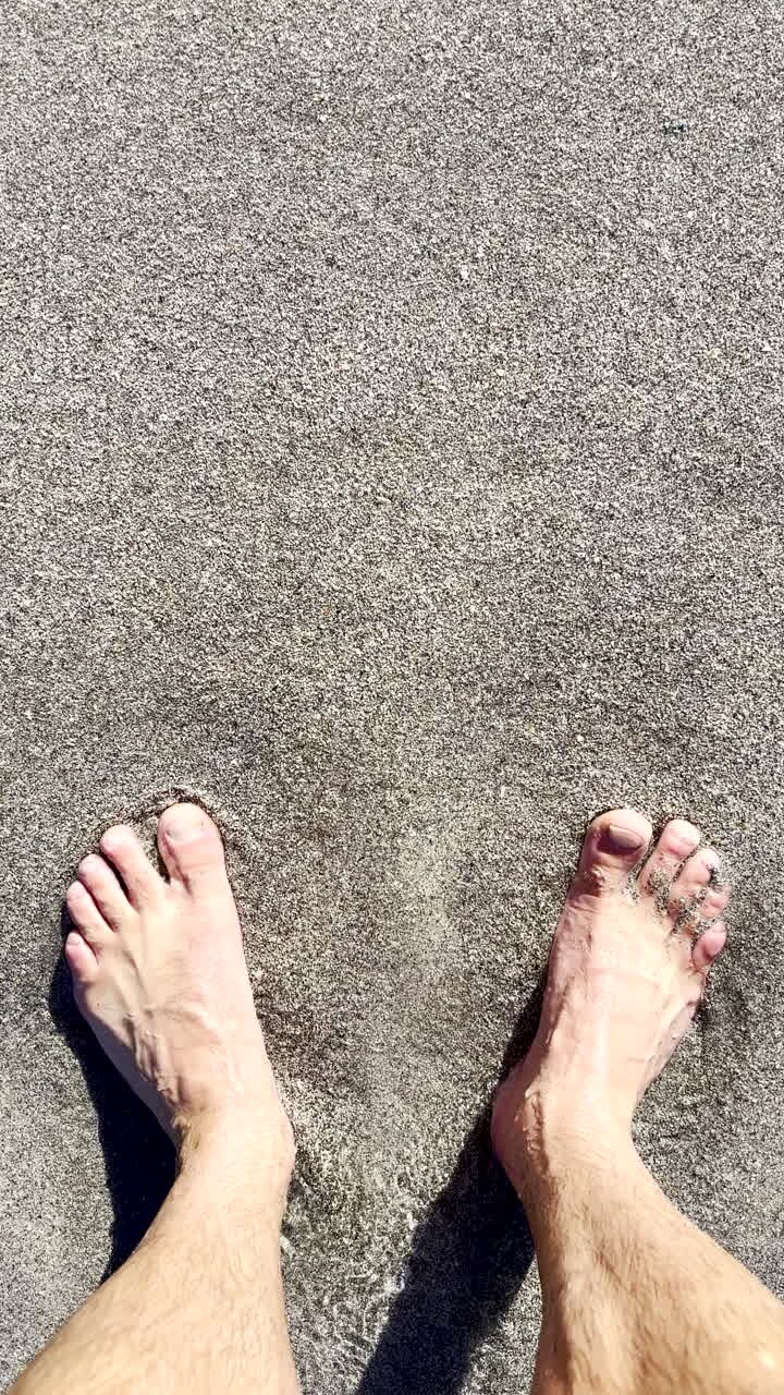 Vista verticale dall'alto di un uomo in piedi sulla spiaggia mentre le onde lambiscono i suoi piedi