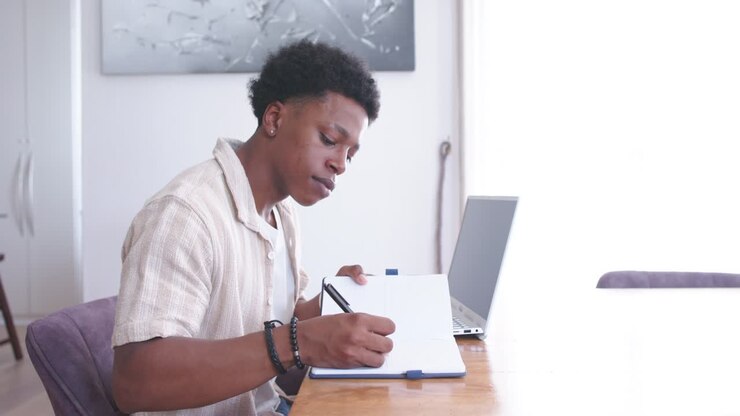 Uomo afroamericano sui vent'anni appoggiato, con la penna sulla pagina, prende appunti al tavolo, dando un'occhiata al laptop