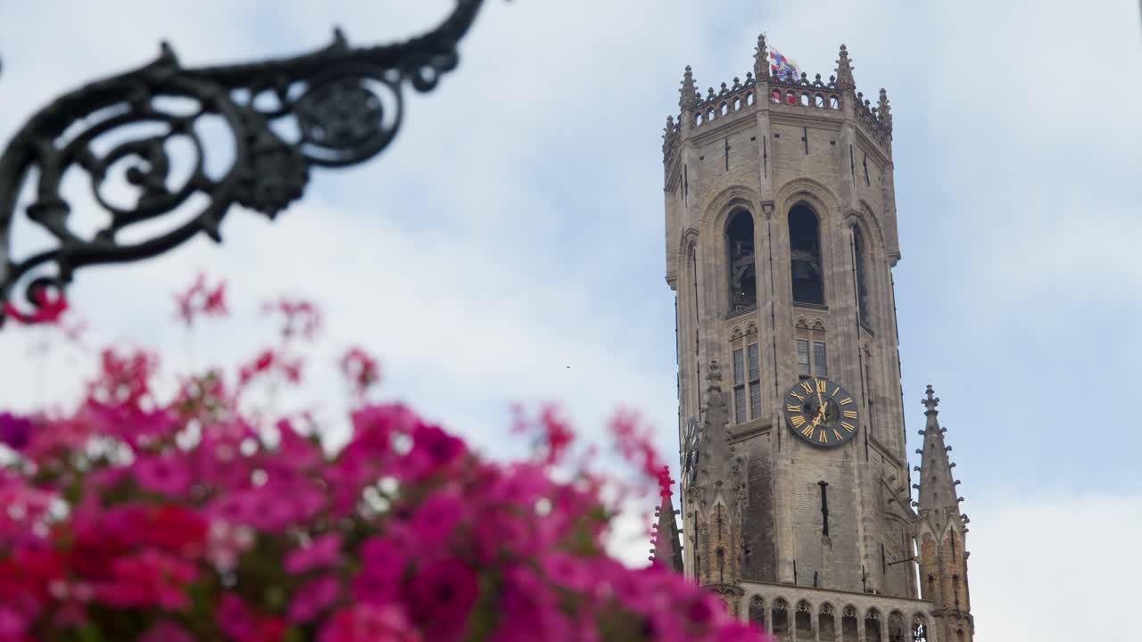 Gothic belfry tower rises above pink flowers, ornate ironwork, and blue sky, slight camera pan