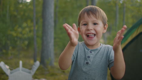 happy cute child boy is clapping and looking at camera at forest camp during weekend family picnic at nature