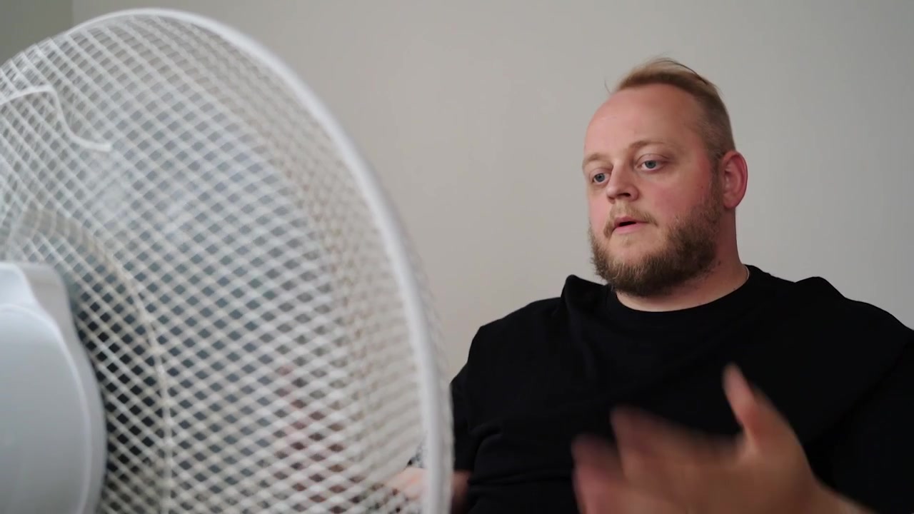 Un uomo sudato con una maglietta nera si china verso un ventilatore elettrico bianco e ne aumenta la velocità, cercando di rinfrescarsi nell'aria calda dell'ambiente