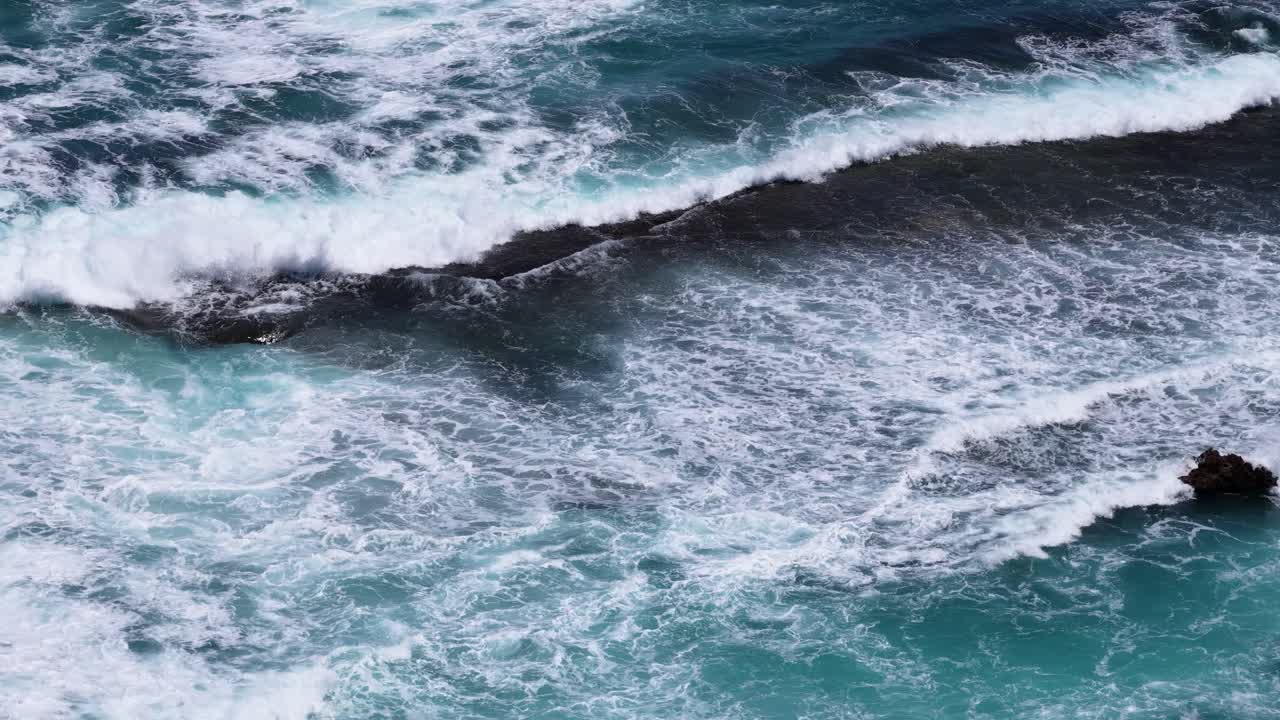 High angle drone footage captures powerful turquoise waves breaking over dark coastal rock formations
