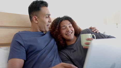 Happy-diverse-gay-male-couple-on-bed,-having-coffee-and-using-laptop-in-the-morning,-slow-motion