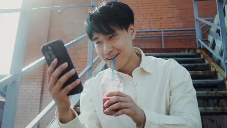 joven usando un teléfono inteligente mientras bebe un batido en las escaleras