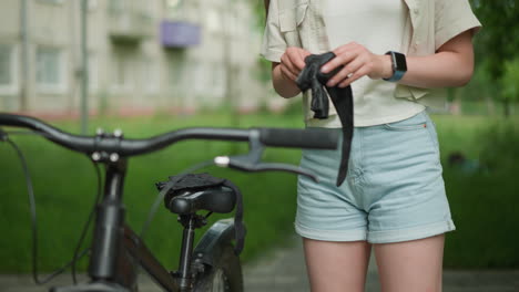 close-up of lady holding black gloves and wearing smart watch on right hand, preparing to wear gloves near her parked bicycle on paved path, with blurred greenery and building in background