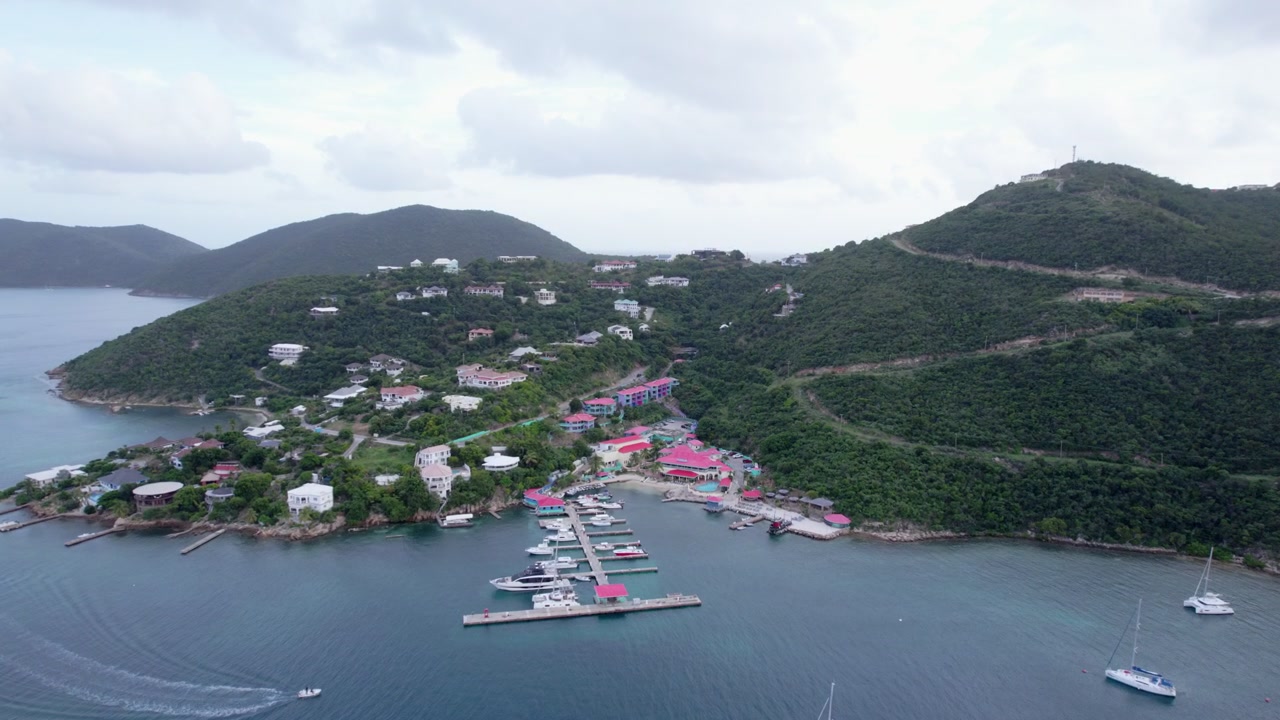 Una veduta aerea di una giornata nuvolosa su Leverick Bay nelle Isole Vergini Britanniche mentre una barca entra nella baia, con la telecamera che si sposta dall'acqua verso il resort, catturando il sereno paesaggio costiero