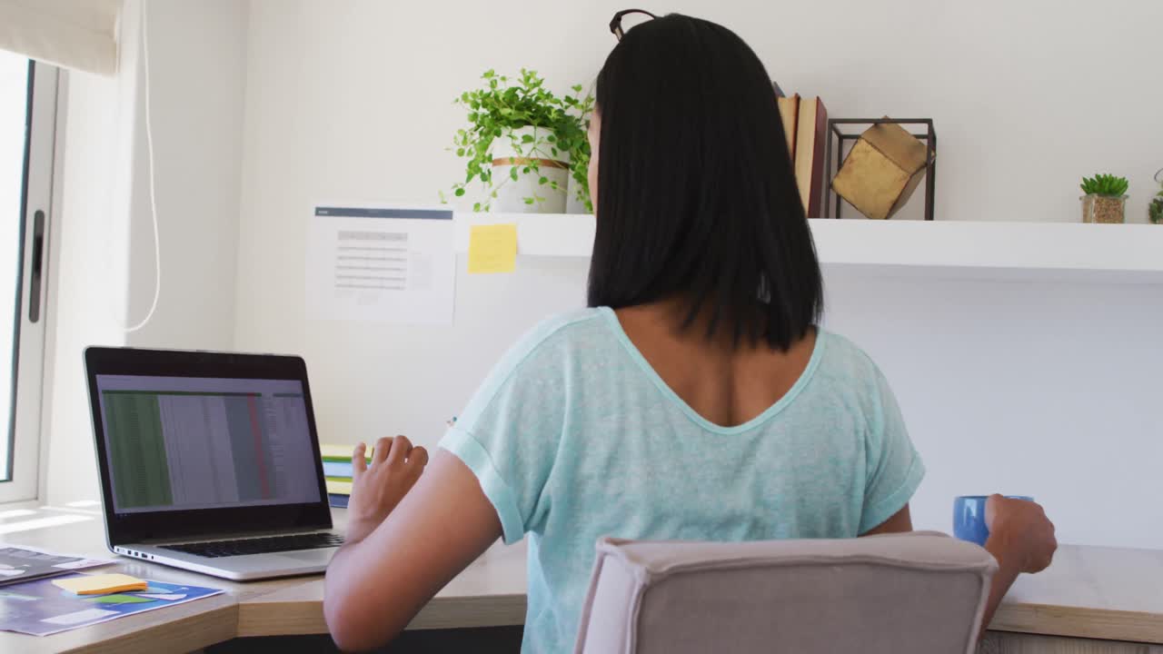 persona de género mixto fluida sentada en un escritorio bebiendo café trabajando desde casa usando una computadora portátil