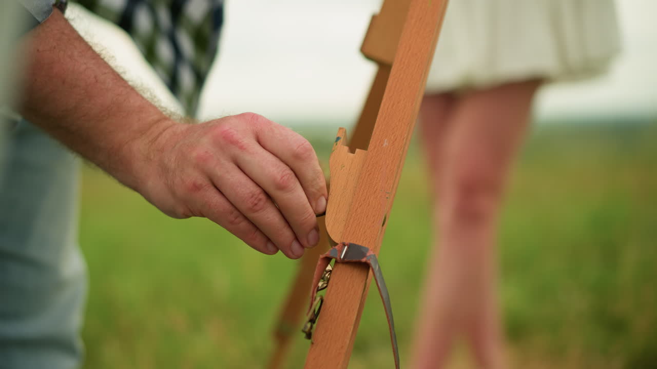 잔디에 있는 야외에서 나무 삼각대를 부드럽게 조정하는 남자의 손의 클로즈업 이미지. 배경에는 여성의 다리가 보이며, 무대에 진정하고 집중된 분위기를 더합니다.