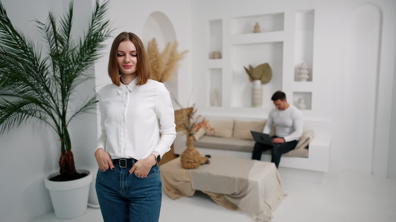 Woman in white shirt and jeans posing in the room in front of camera. Business lady portrait. Man sits at backdrop using laptop.