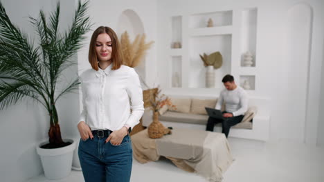 woman in white shirt and jeans posing in the room in front of camera. business lady portrait. man sits at backdrop using laptop.