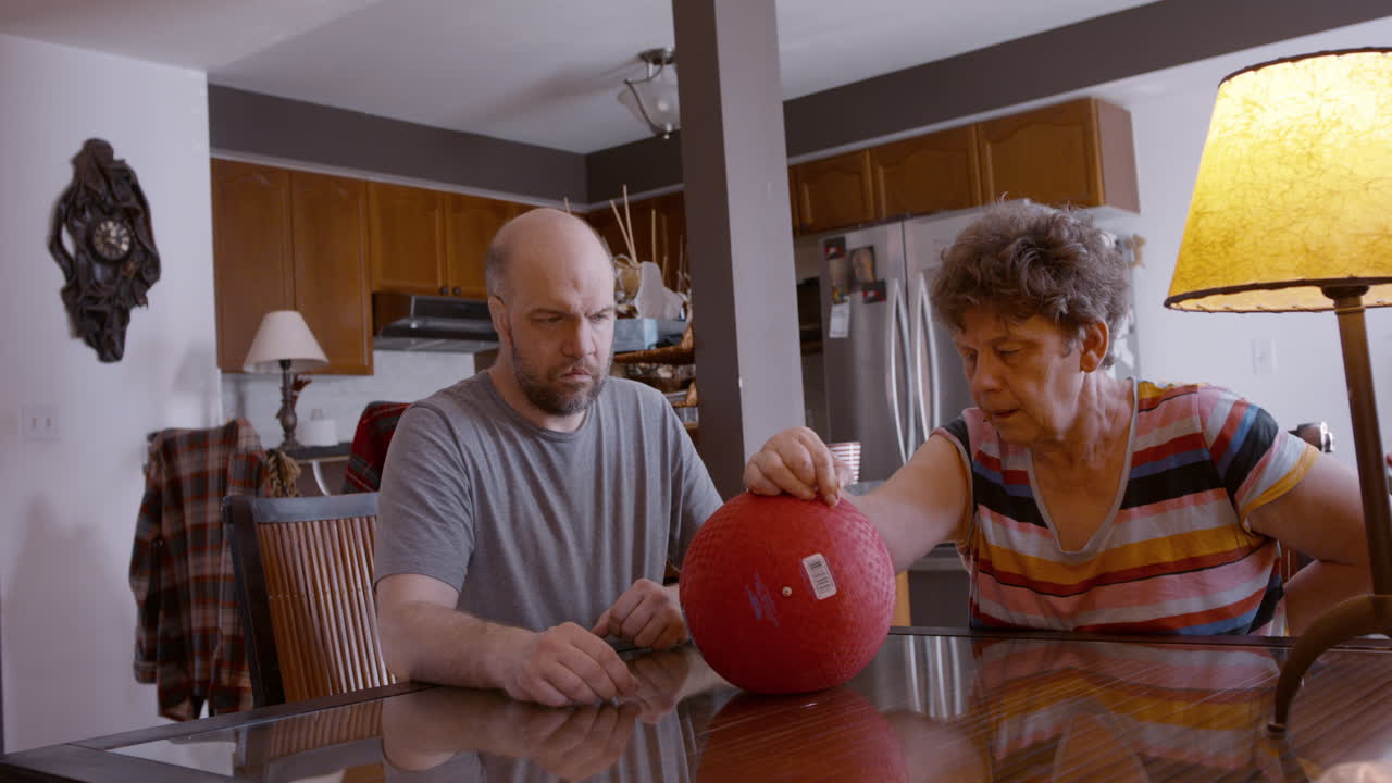 Onlooking middle-aged male caregiver gently assists the hemiplegic right hand ball therapy of an elderly female stroke survivor, seated across from him on glass top kitchen table at home. Medium shot.