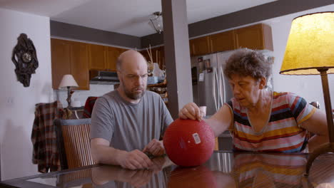 onlooking middle-aged male caregiver gently assists the hemiplegic right hand ball therapy of an elderly female stroke survivor, seated across from him on glass top kitchen table at home. medium shot.