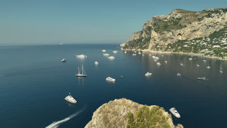 Backwards-drone-dolley-shot-of-luxury-yachts-anchored-at-Capri-rocks-in-Italy-on-a-sunny-day