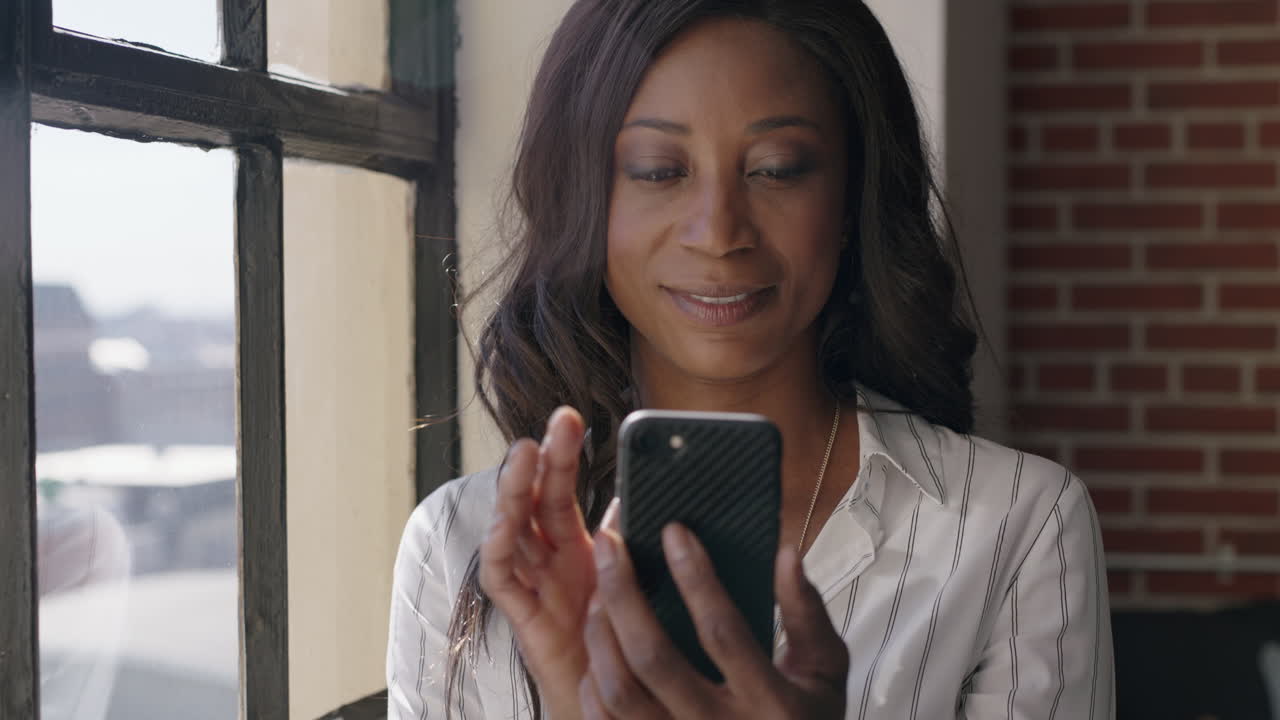 ritratto di una donna afroamericana felice che usa lo smartphone, beve caffè a casa, naviga i messaggi dei social media, gode di una mattinata rilassata, guarda fuori dalla finestra, invia messaggi sul cellulare