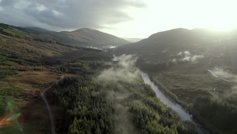 Drohnenaufnahmen-Aus-Der-Luft,-Die-Langsam-Fliegen,-Hoch-über-Einem-Fluss,-Einer-Straße-Und-Einem-Nadelwald,-Während-Tief-Hängende-Wolken-Die-Baumkronen-Umarmen-Und-Die-Sonne-Hinter-Bergen-Untergeht