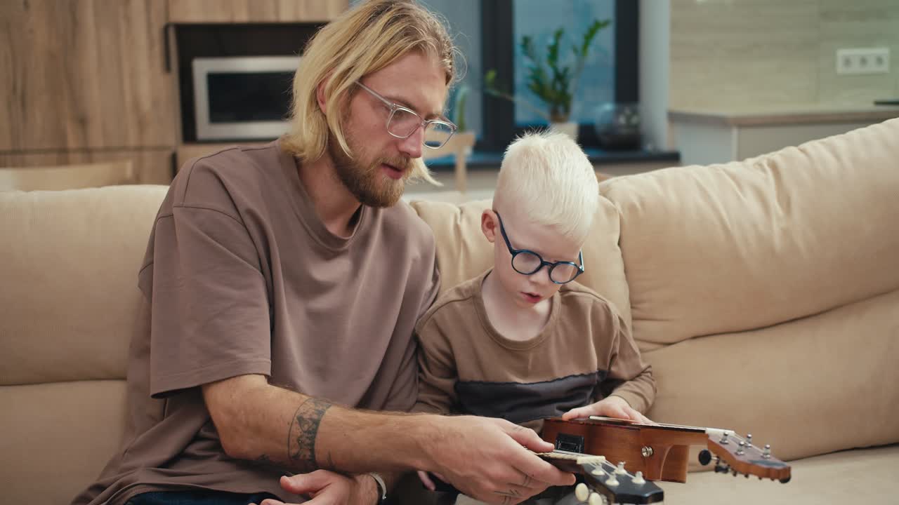 A blond man in glasses with a beard tells his little son an albino boy with white hair in blue glasses how a ukulele works and points to the strings while learning music in a modern apartment