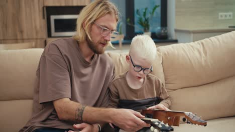 Ein-Blonder-Mann-Mit-Brille-Und-Bart-Erklärt-Seinem-Kleinen-Sohn,-Einem-Albino-Jungen-Mit-Weißen-Haaren-Und-Blauer-Brille,-Wie-Eine-Ukulele-Funktioniert,-Und-Zeigt-Auf-Die-Saiten,-Während-Er-In-Einer-Modernen-Wohnung-Musik-Lernt