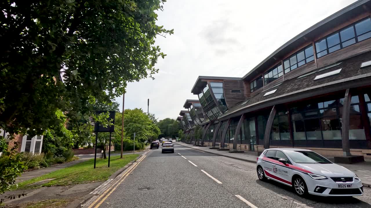 A vehicle travels along a tree-lined street with historic brick houses and a contemporary university building under daylight, captured in smooth forward motion