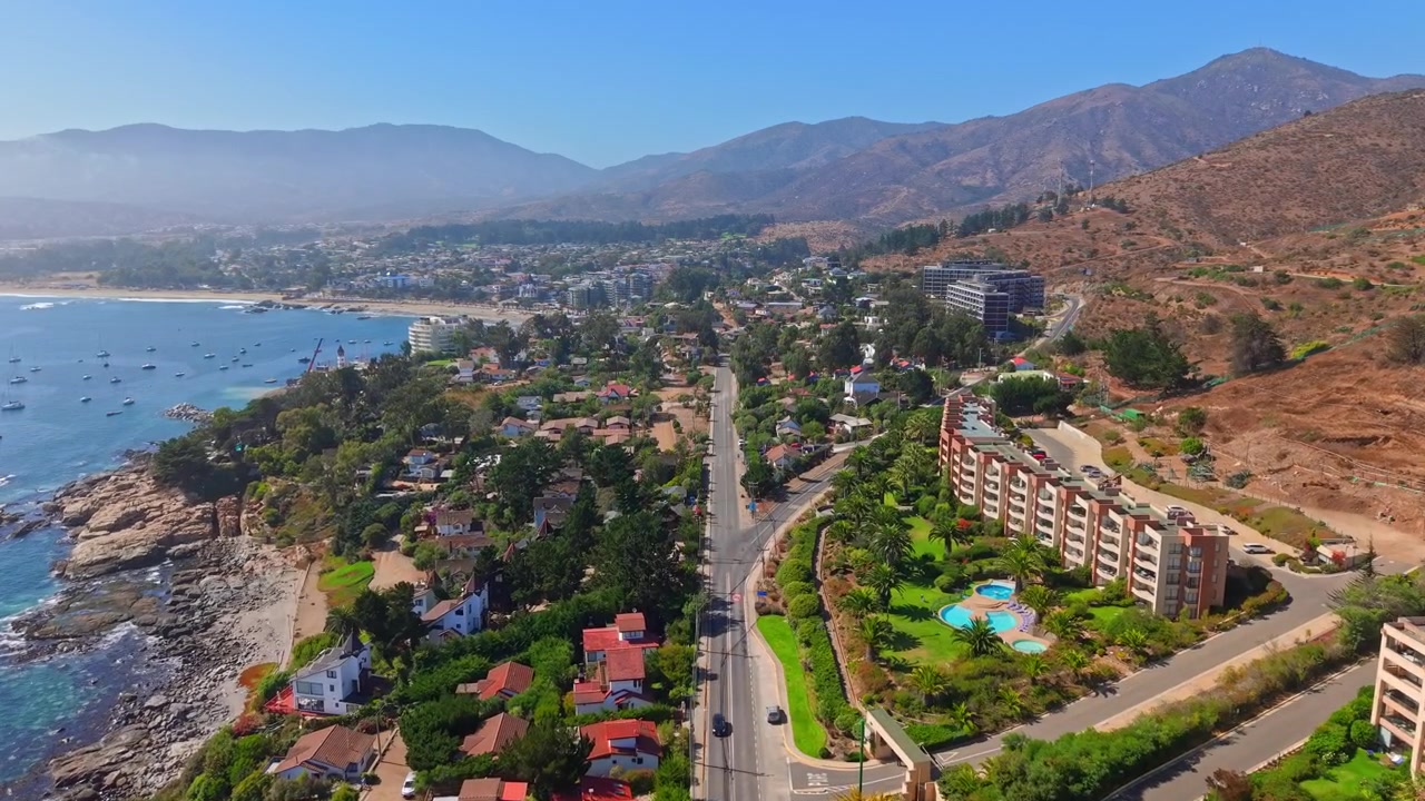 Vista aérea de la entrada a Papudo, Chile, mostrando casas de lujo frente al mar y edificios residenciales en una mañana neblinosa, con yates en la playa