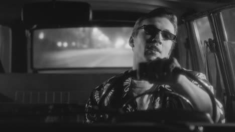 black and white shot of man in sunglasses driving vintage car at night