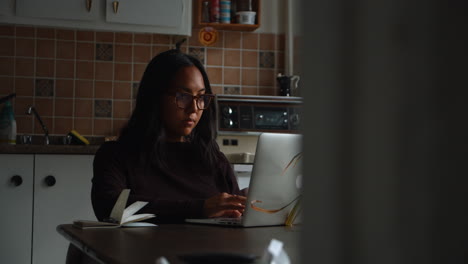 a young asian woman is sitting at her dinner table as she uses her laptop computer to work from home