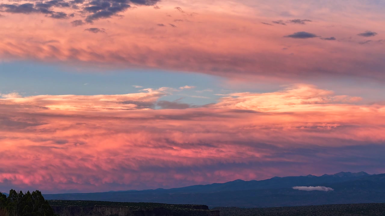 4K UHD drone timelapse of a surreal, thick curtain of clouds drifting over a dark mountain horizon. Vibrant sunset colors transition through the sky, illuminating the textured cloud layers.