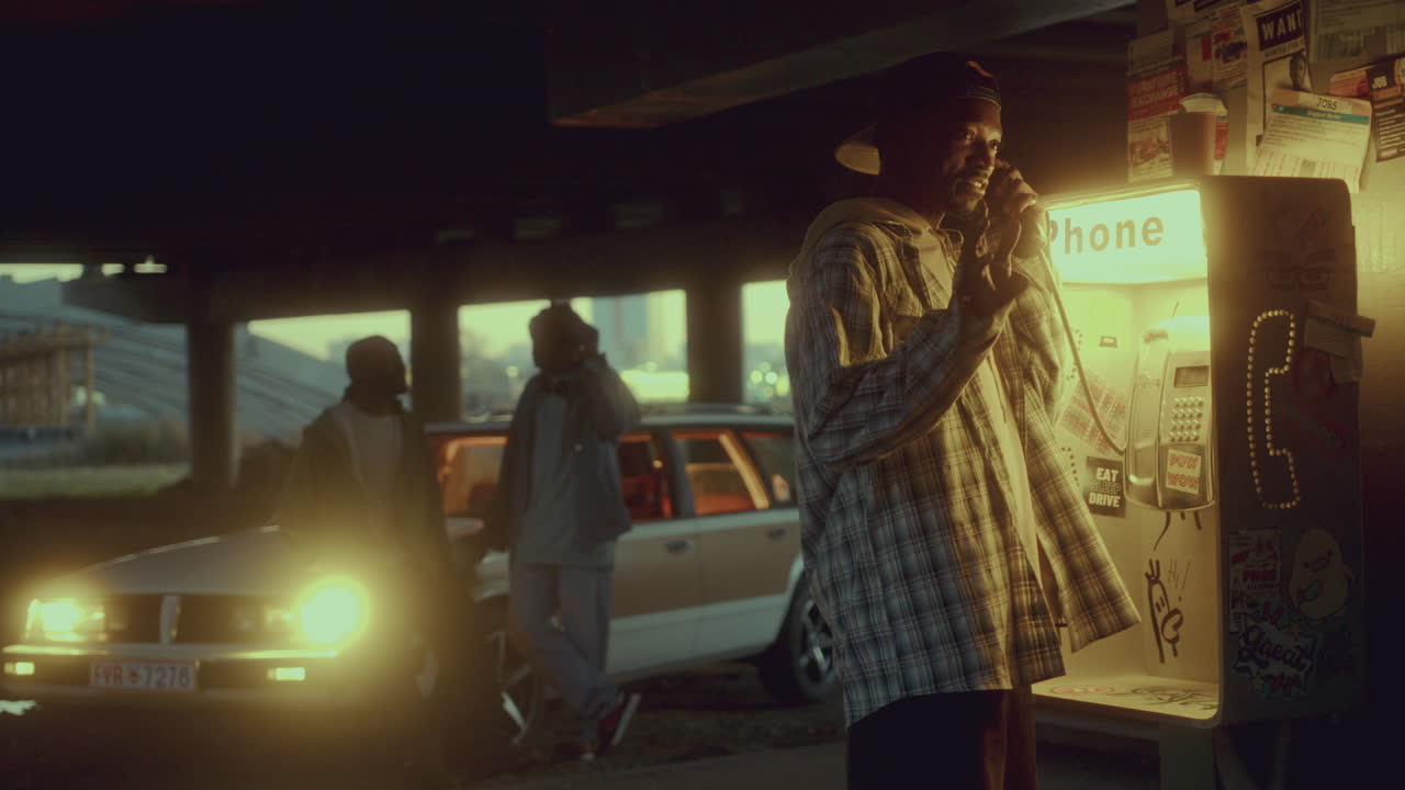 흑인 남자가 도시 지역의 희미하게 조명 된 오버브리스 에서 유료 전화로 이야기하고 있습니다.