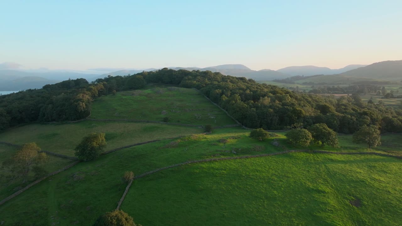 Woodland covered hilltop at sunrise with camera rise showing lake and distant mountains. Summer. Windermere, Cumbria, UK