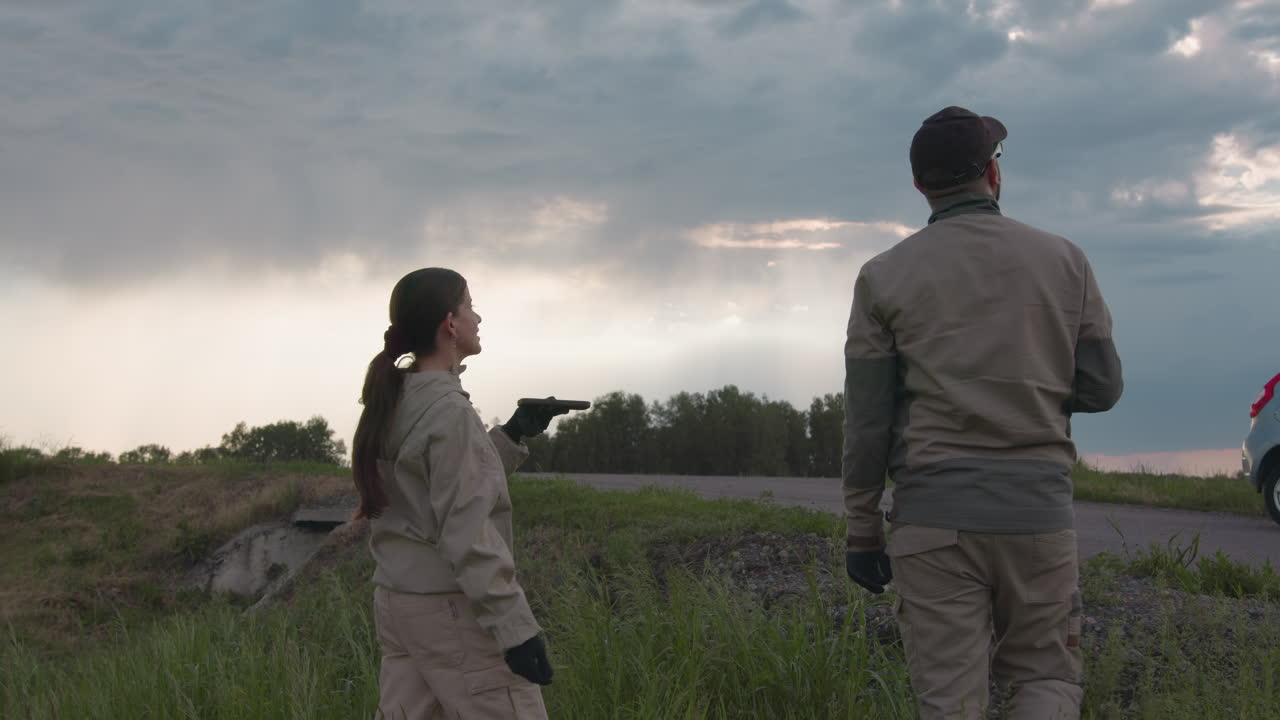 Couple in matching uniform stands on grassy roadside under moody sky, woman releases paraglider balloon while holding handheld device, both looking upward as soft light breaks through layered clouds