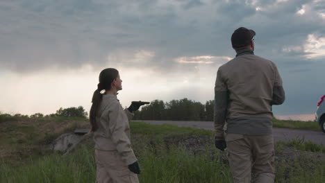 couple in matching uniform stands on grassy roadside under moody sky, woman releases paraglider balloon while holding handheld device, both looking upward as soft light breaks through layered clouds