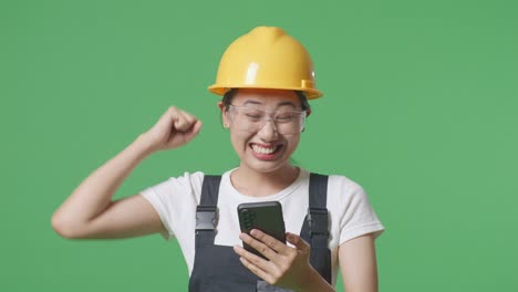 close up of asian woman worker wearing goggles and safety helmet looking at smartphone then screaming goal celebrating while standing in the green screen background studio